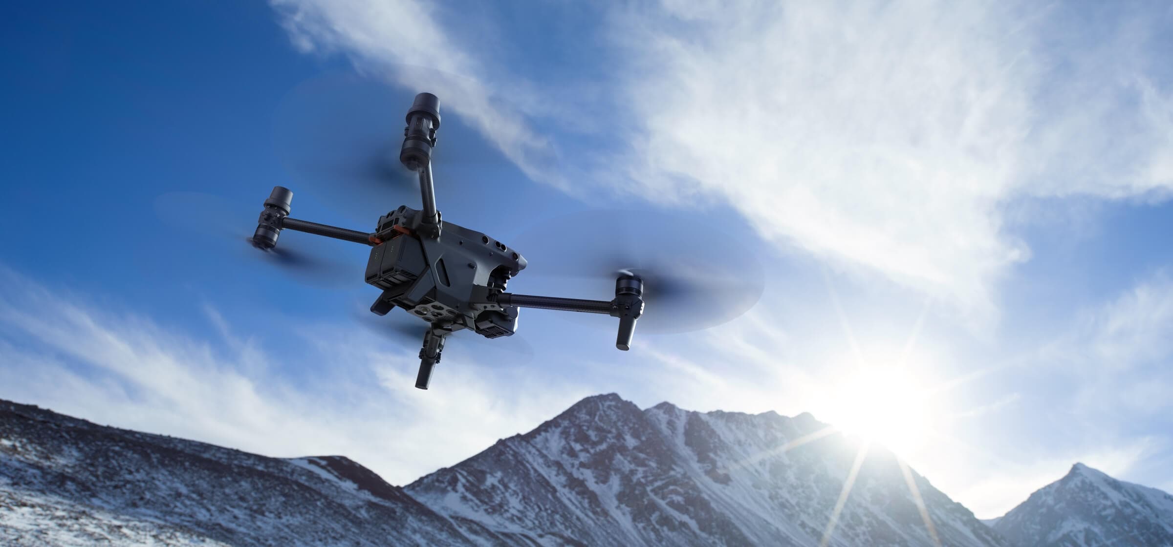 DJI Matrice 30 u letu iznad snjeznih planinskih vrhova, propeleri u pokretu, sunce probija kroz oblake iza drona